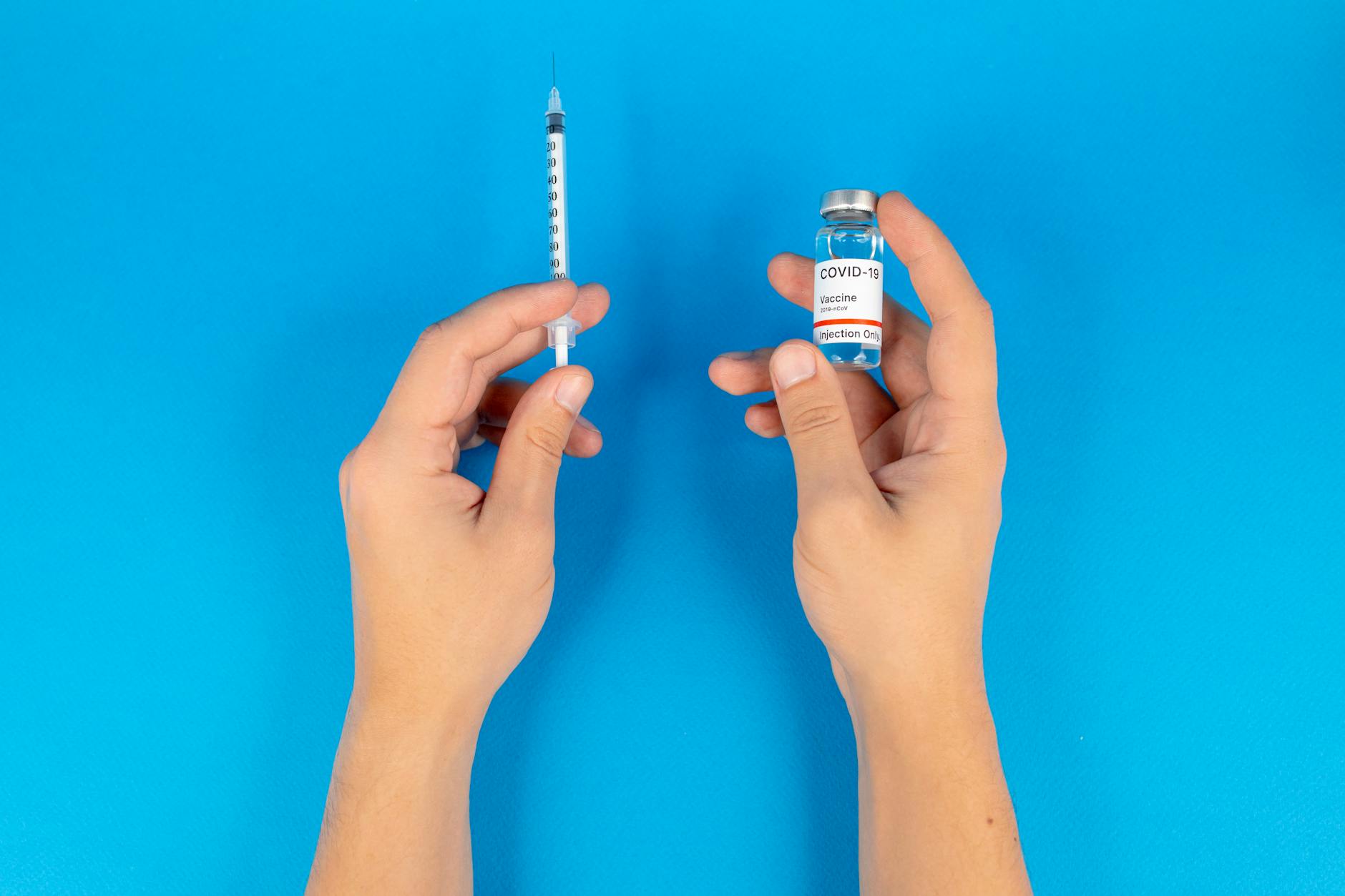 Close-up of hands holding a COVID-19 vaccine vial and syringe against a blue background.